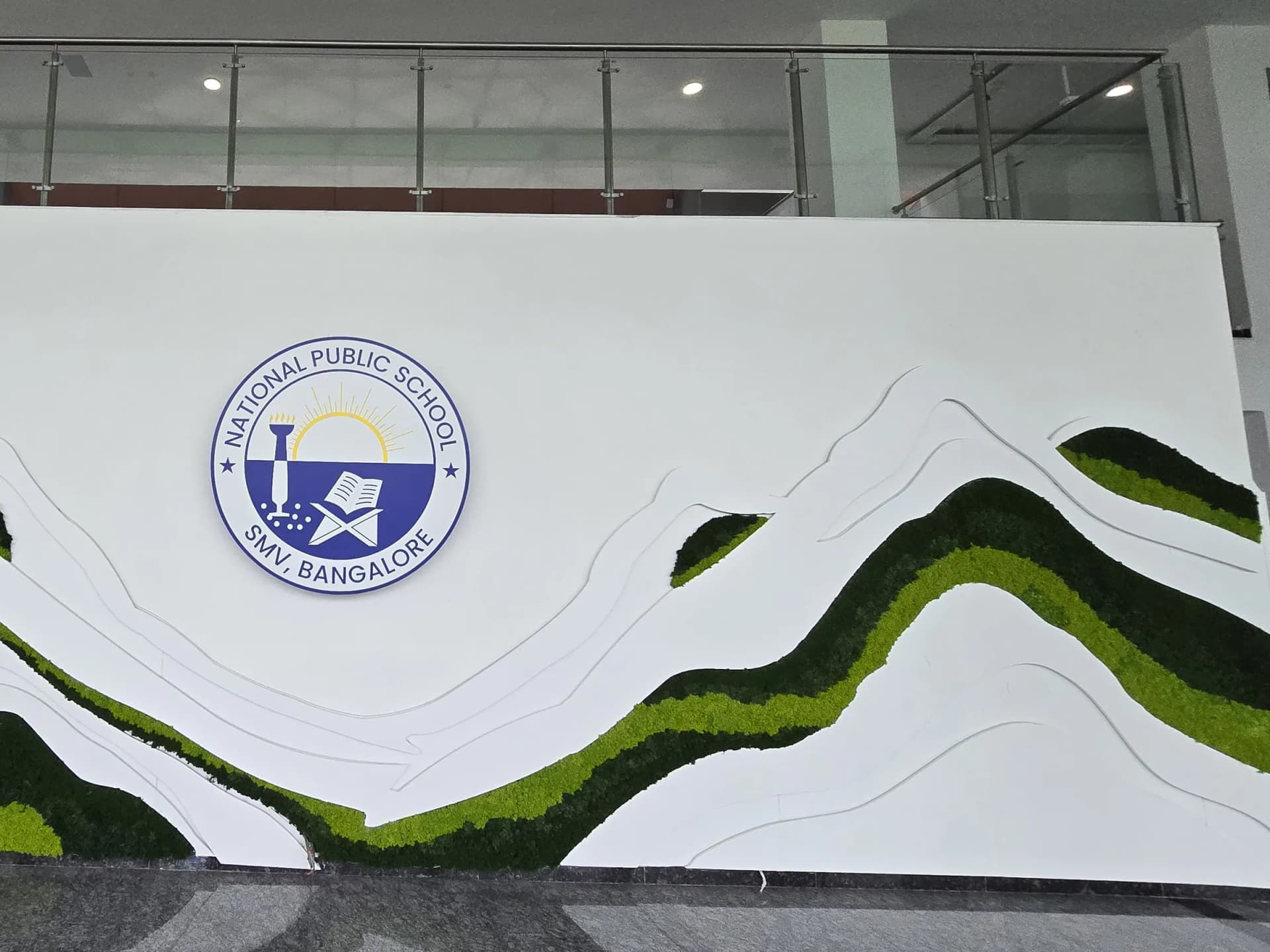 Close view of the National Public School SMV Bangalore crest wall, showing the layered white relief and the two greens of preserved moss
