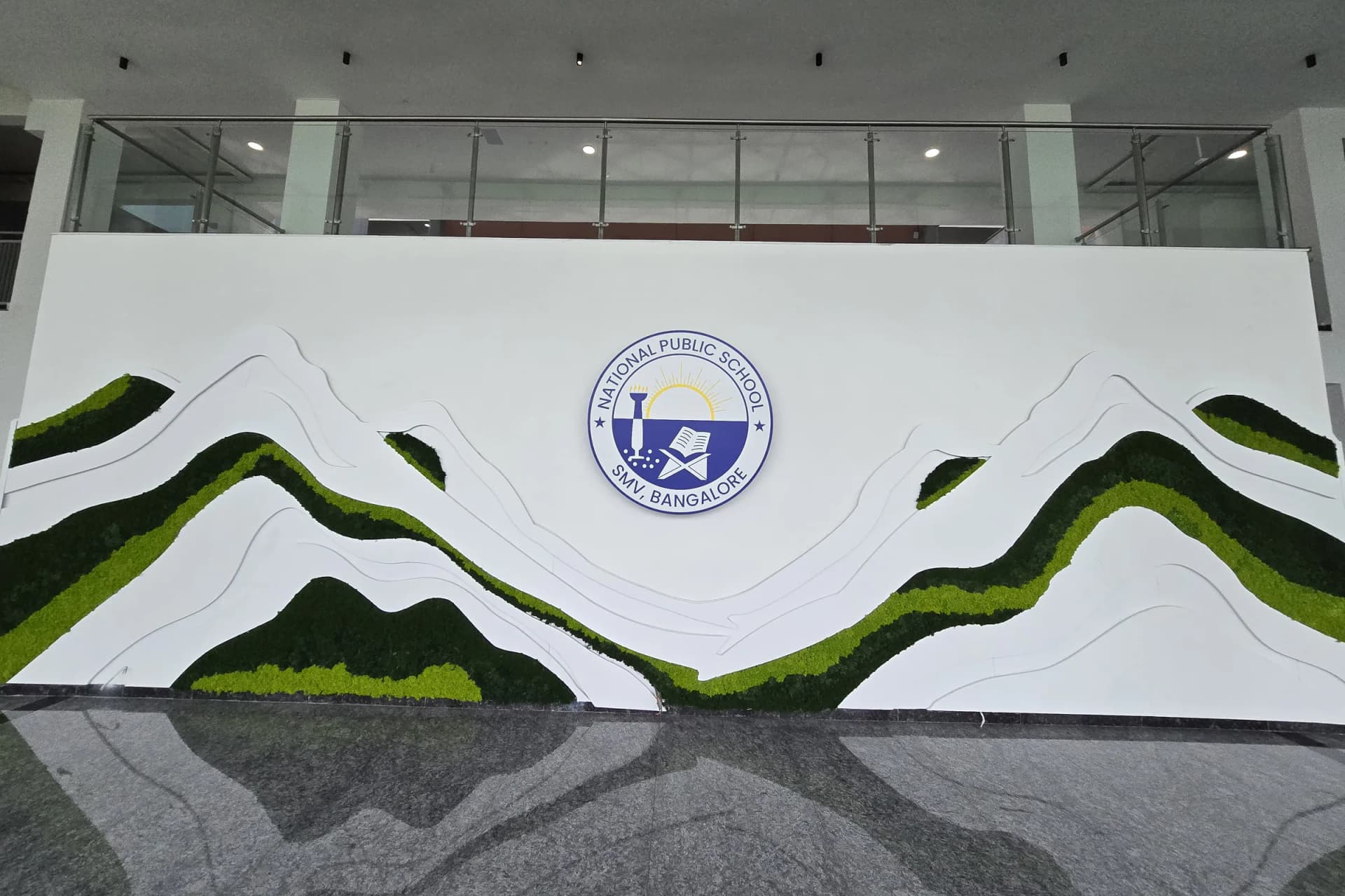 Reception feature wall at National Public School, SMV Bangalore: layered white relief hills edged with preserved moss in two greens, the school crest at the centre