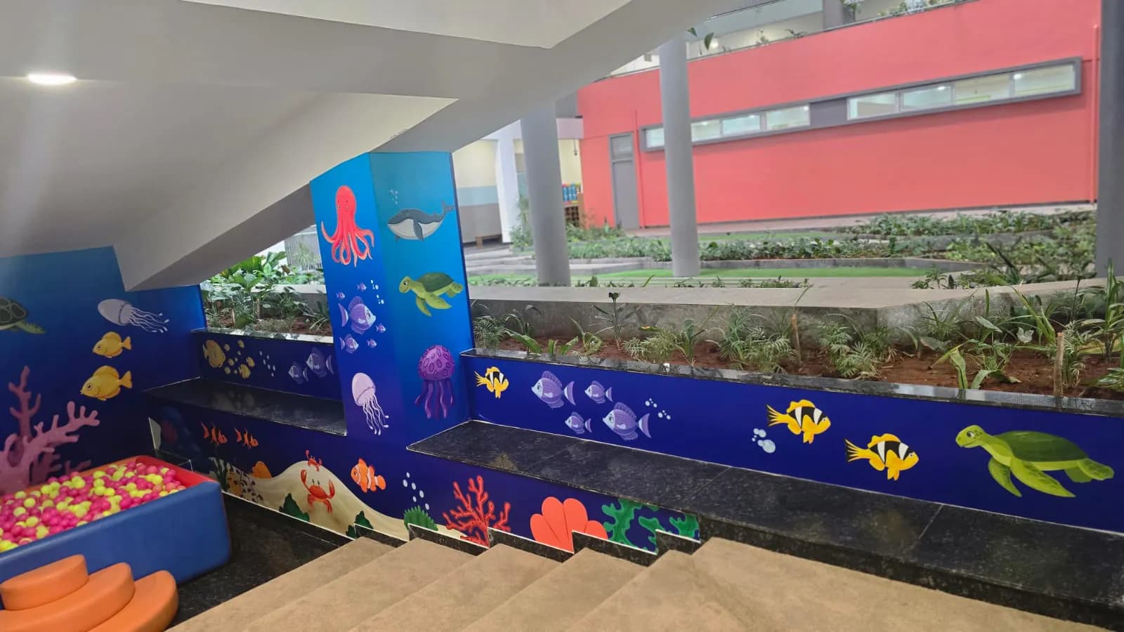 Underwater graphics wrapping the pillars, planter faces and step risers beside a ball pit, with the school courtyard behind