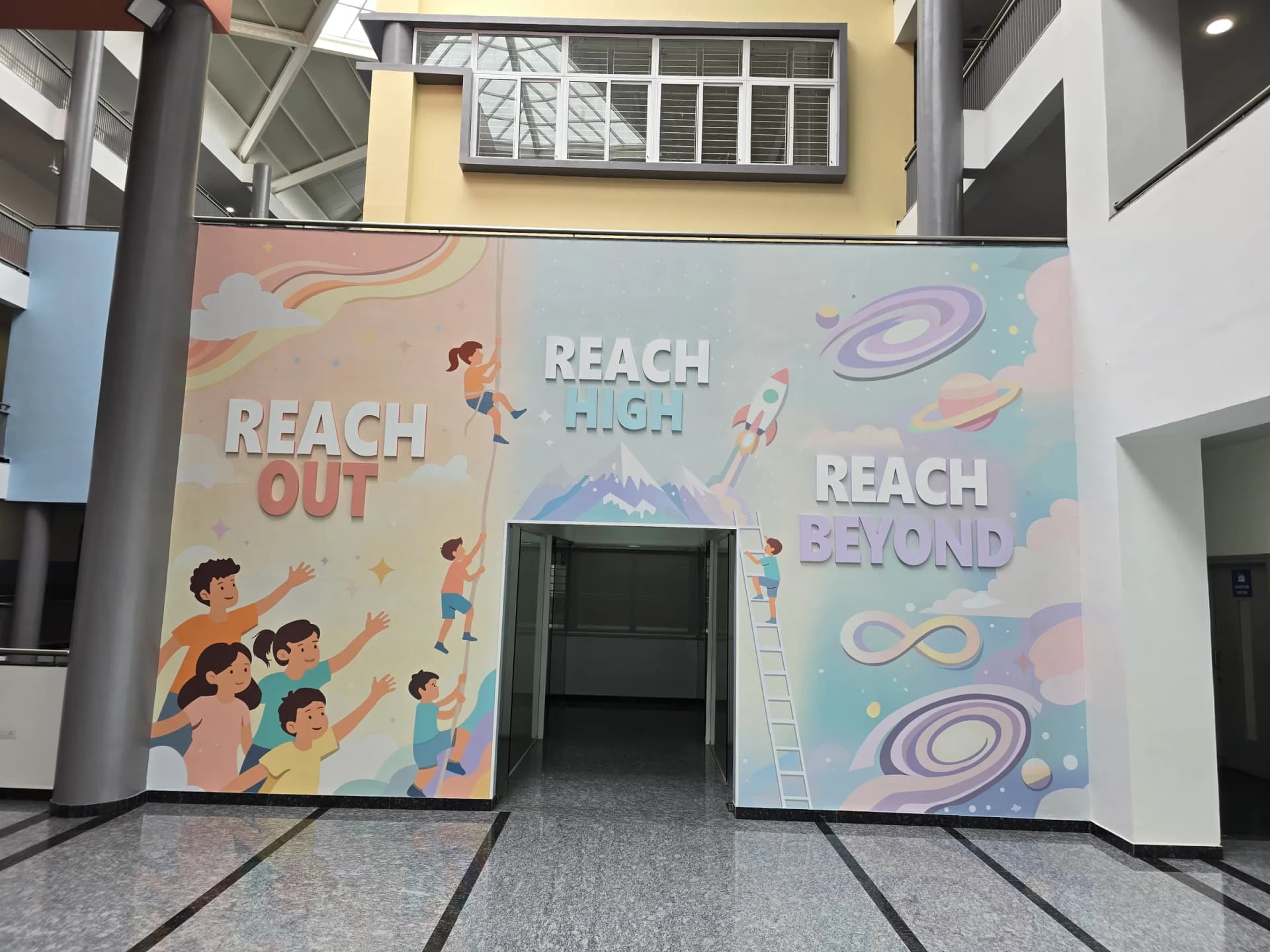 Atrium wall graphic around a doorway: children climbing a rope and a ladder toward a rocket and planets, with Reach Out, Reach High and Reach Beyond in dimensional letters
