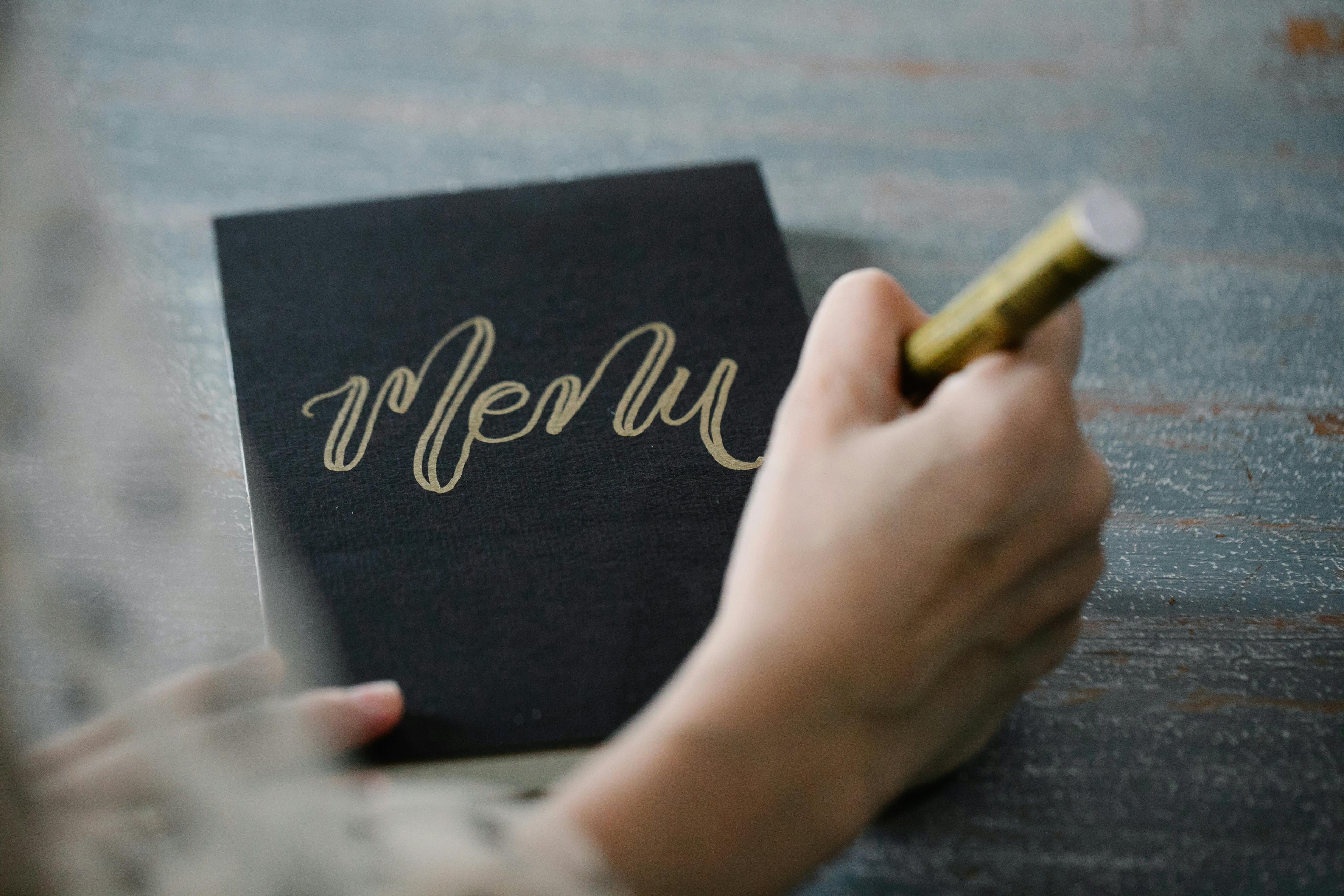 Close-up of a hand holding a gold marker writing on a black menu card on a rustic wooden table